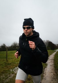 Homme portant une veste noire, un bonnet et des lunettes de soleil courant sur un chemin de campagne boueux sous un ciel couvert.