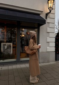 Manteau en laine beige avec ceinture à nouer, bonnet en tricot beige, et sac épaule en cuir marron. Debout sur un trottoir en pierre près d'une vitrine de magasin.