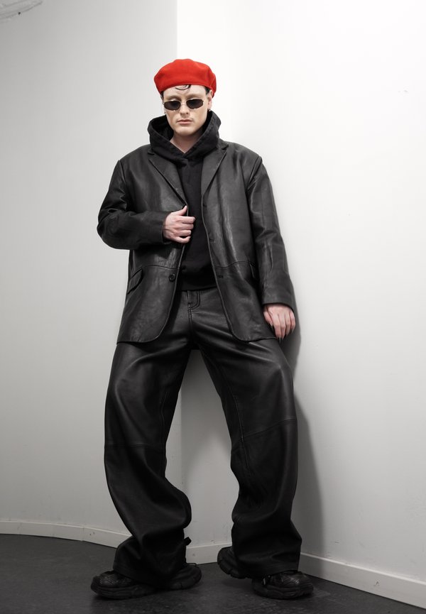 Persona, kura valkā sarkanu bereti, melnas saulesbrilles, pārmērīgi lielu melnu ādas jaku un bikses, stāv pret balto sienu ar nopietnu izteiksmi.