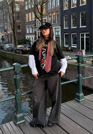 Camiseta gráfica negra con diseño bordado de serpiente y flores, superpuesta a una camisa de manga larga blanca. Pantalones anchos gris oscuro y zapatos negros.