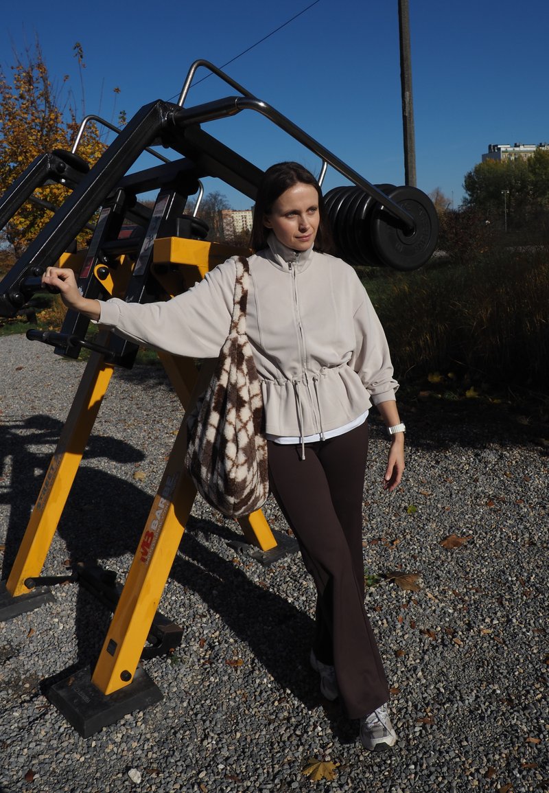 Light beige jacket with a cinched waist, brown pants, and patterned tote bag. Outdoor gym equipment and gravel surface in background.