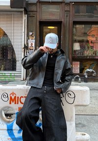 Svart skinnjakke, tettsittende svart t-skjorte, oversized mørke denimbukser med knapper og lys blå caps med hvit logo. Urban gateomgivelse.