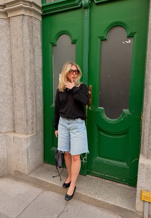 Mujer con pelo rubio y gafas de sol está delante de grandes puertas dobles verdes, lleva una camiseta negra, pantalones cortos de mezclilla claros y zapatos planos negros.