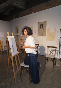 Une femme portant un t-shirt blanc et un jean en denim à jambes larges est à genoux à côté d'un chevalet, esquissant des dessins affichés sur le mur dans un cadre de studio d'art.