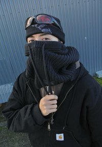 Črna pletena ruta z vsebnostjo teksturiranega vzorca, nosi se visoko okoli vratu. Črna pula in kapa s sončnimi očali. Siva ozadje.