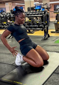 Tapis de fitness noir avec une personne en tenue sportive noire à genoux, effectuant un exercice. Baskets blanches, salle de sport en arrière-plan.