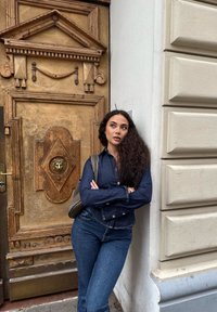 Jeanshemden und Jeans in mittelblau, stehend gegen eine strukturierte Holztür mit aufwendigen Details und einem Löwenkopfklopfer.