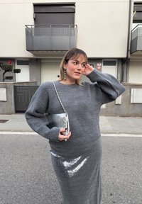 Γυναίκα που στέκεται στον δρόμο με γκρι πουλόβερ και λαμπερή φούστα με παγιέτες, κρατώντας ασημένια τσάντα, αγγίζοντας τα μαλλιά της, με κτίρια και μπαλκόνια στο παρασκήνιο.