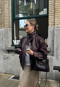 Jachetă din piele vișinie, cămașă albă și fustă bej. Ține în mână o băutură verde și o geantă neagră țesută cu un detaliu auriu.