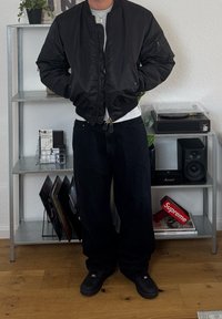 Fekete bombázó dzsekit, szürke pólót és fekete bő szárú nadrágot viselő személy áll egy fapadlón egy fém polc mellett, amely lemezeket és audio berendezéseket tartalmaz.