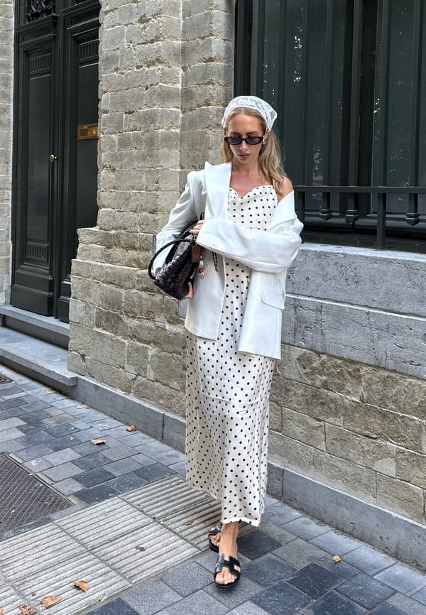 Blazer oversize bianco, vestito a pois in crema e nero, sandali neri e borsa nera. Muro di pietra texture sullo sfondo.