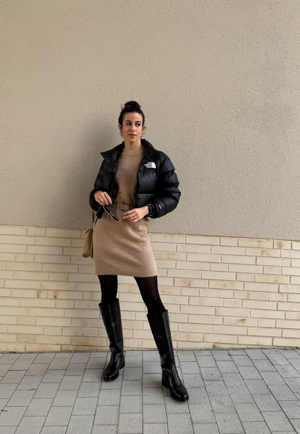 Μαύρο puffer μπουφάν πάνω από μια μπεζ πλεκτή φόρεμα, συνδυασμένο με μαύρες δερμάτινες μπότες μέχρι το μηρό και καφέ ζώνη, καθώς και μπεζ τσάντα.