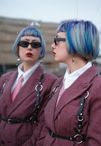 Deux personnes avec des cheveux pastel bleu et violet à la hauteur du menton portant des lunettes de soleil noires. Elles sont habillées en costumes à chevrons roses avec des bretelles et des chemises blanches.