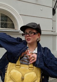 Sac en cuir jaune avec double poignée, veste en denim à manches surdimensionnées, chemise rayée en dessous, plusieurs anneaux et lunettes épaisses.