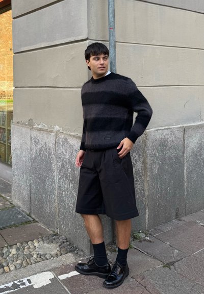 Suéter de rayas negras en material peludo, combinado con pantalones cortos negros holgados y zapatos negros. De pie contra una pared gris claro.