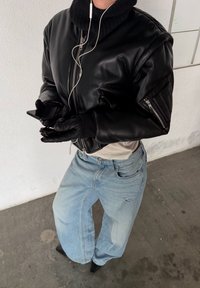 Veste bombardier en cuir noir avec un col montant, associée à un jean bleu clair à jambes larges. La silhouette tient un smartphone et porte des gants en cuir.