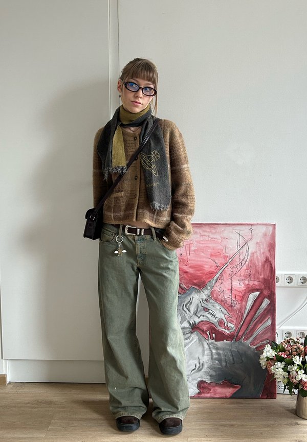 Νέος με γυαλιά, που φοράει καφέ πουλόβερ, πράσινο κασκόλ και φαρδιά πράσινα παντελόνια, στέκεται δίπλα σε μια ζωγραφιά με μονόκερο και ένα βάζο με λουλούδια.