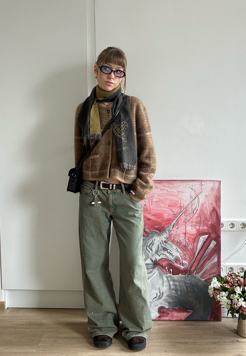 Eine junge Person mit Brille, die einen braunen Pullover, einen grünen Schal und weite grüne Hosen trägt, steht neben einem Einhorn-Gemälde und einer Vase mit Blumen.