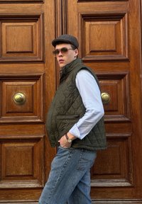 Gilet trapuntato verde oliva con colletto alto, tasche laterali, indossato sopra una camicia azzurro chiaro, abbinato a jeans di denim e un cappellino grigio. Porta di legno sullo sfondo.