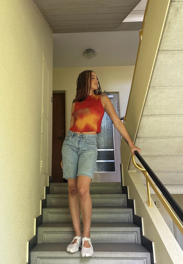 Jonge vrouw met vlechten, gekleed in een rood-oranje mouwloos topje en spijkershort, staat op een binnentrap en houdt een zwart handvat vast.