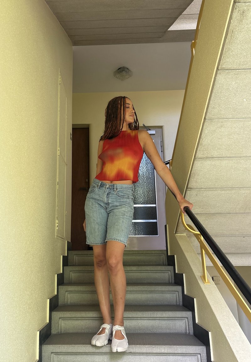 Jeune femme avec des nattes portant un haut sans manches rouge-orange et un short en denim, debout sur des escaliers intérieurs tenant une rampe noire.