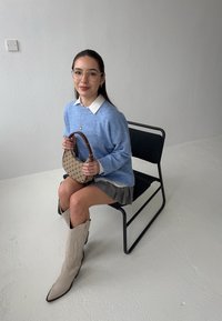 Světle modrý pletený svetr, bílá košilová blůza, vzorovaná sukně, béžové kovbojské boty, držící malou hnědo-krémovou kabelku, sedící na černé židli.