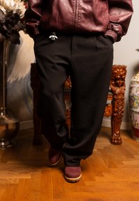 Personne portant une veste en cuir bordeaux, un pantalon noir et des bottes bordeaux, avec un petit porte-clés accroché à la poche, debout sur un sol en bois.