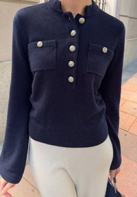 Kvinne iført en marineblå strikket topp med sølvknapper og lapplommer, kombinert med hvite bukser og holder en svart håndveske.