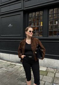Veste en daim marron sur un haut noir, associée à un pantalon court noir. Les accessoires comprennent des lunettes de soleil rondes et un petit sac à main noir.