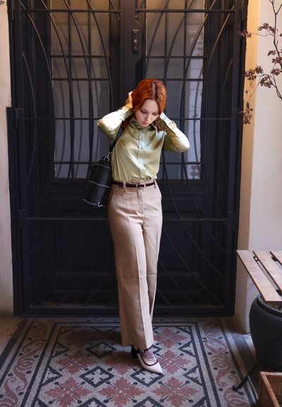 Mujer pelirroja se acomoda el cabello, vestida con blusa verde, pantalones beige y zapatos puntiagudos, de pie sobre un suelo de baldosas con patrón junto a una puerta de hierro negra.