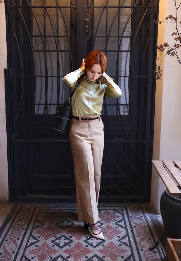 Rød-håret kvinne justerer håret, iført grønn bluse, beige bukser og spisse sko, stående på mønstret flislagt gulv ved en svart jernport.