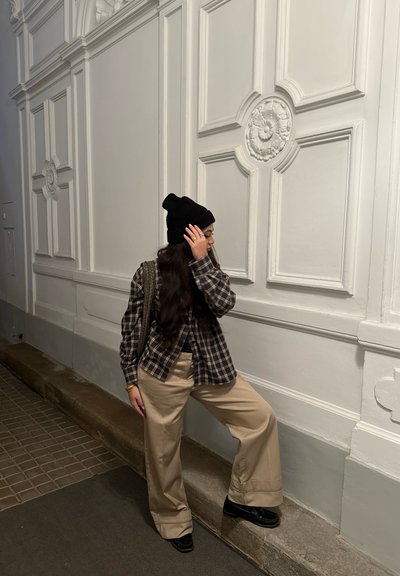 Persona con gorro negro, camisa de cuadros, pantalones beige y zapatos negros, de pie con un pie levantado sobre un borde frente a una pared blanca ornamentada.