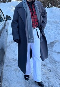 Manteau gris, chemise à carreaux rouge et noir, pantalon blanc, écharpe grise, gants noirs. Debout sur un sol enneigé près d'une voiture.