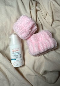 Avene Cleanance vochtinbrenger in een witte fles met rode accenten, naast twee roze pluche washandjes met een geribbelde textuur, op een crème achtergrond.