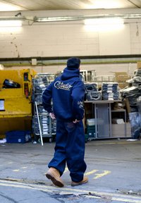 Homme portant un sweat à capuche bleu marine avec le texte « Studio » et un jean ample, marchant dans un entrepôt industriel rempli de marchandises empilées et de palettes.