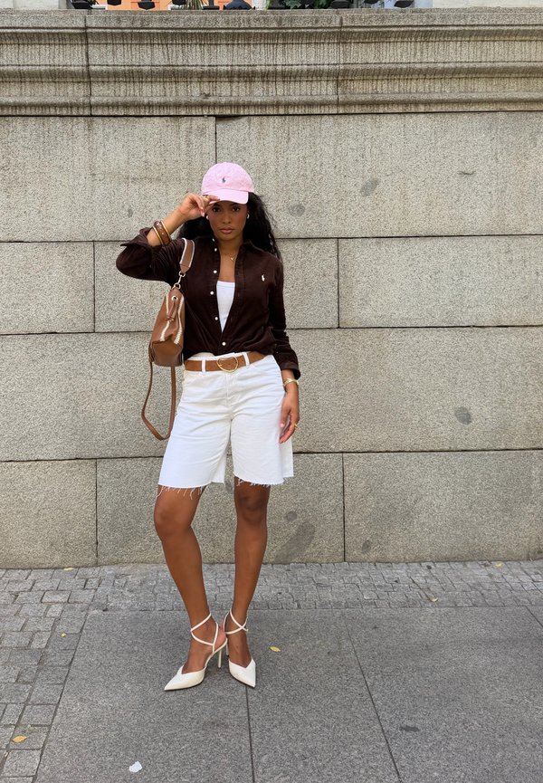 Mujer de pie contra una pared de piedra, vestida con una camisa marrón, pantalones cortos blancos, tacones blancos, gorra rosa y llevando un bolso marrón en el hombro.