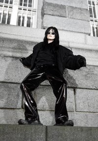 Sort polstret jakke over en mørk top, kombineret med sorte viddebenede bukser med høj glans. Fodtøjet er mørkt og robust, hvilket komplimenterer tøjets tekstur.