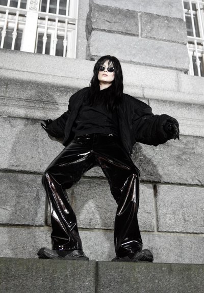 Chaqueta acolchada negra sobre una camiseta oscura, combinada con pantalones anchos negros de alto brillo. El calzado es oscuro y robusto, complementando la textura del conjunto.