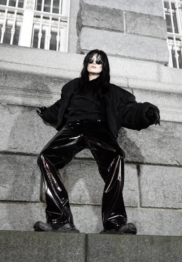 Fekete bélelt dzseki sötét felső fölött, fényes fekete, bőszárú nadrággal párosítva. A lábbeli sötét és masszív, kiegészítve a ruha textúráját.