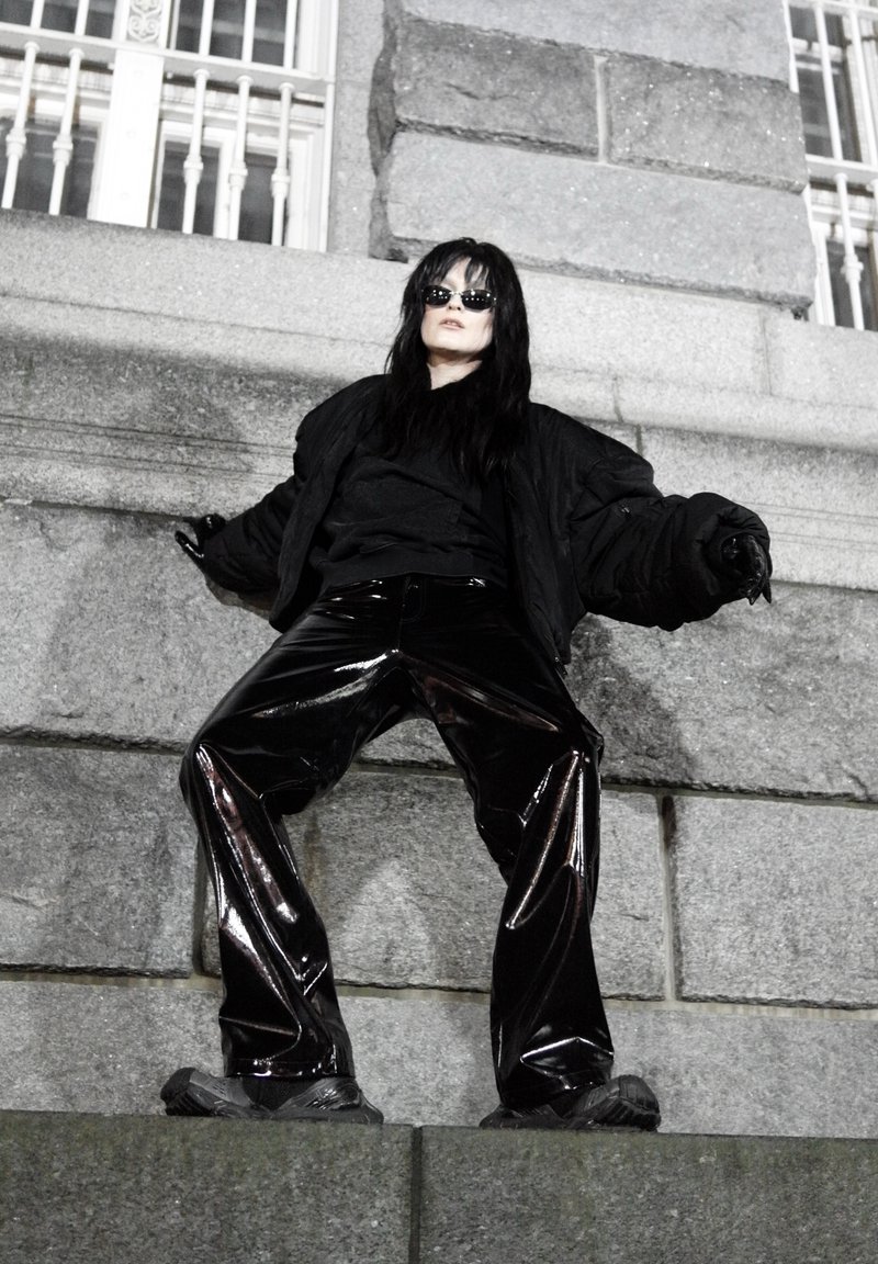 Veste matelassée noire portée sur un haut foncé, assortie à un pantalon noir large à fort éclat. Les chaussures sont sombres et épaisses, complétant la texture de la tenue.