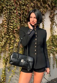 Crna vojnog stila jakna s zlatnim dugmadi, dugim rukavima i plavim manžetama; u kombinaciji s teksturiranom crnom torbom i kratkim hlačama. Pozadina s zelenim lišćem.