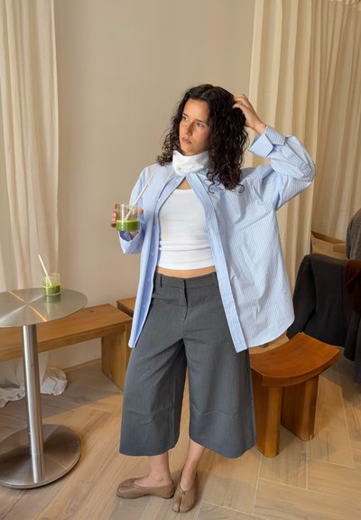 Mujer con cabello rizado y oscuro que lleva una camiseta corta blanca, una camisa de rayas azules y pantalones culottes grises, sosteniendo una bebida verde con pajita en interiores.