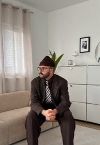 Istuv inimene kannab pruuni ruudulist ülikonda ja lipsu, pruuni beani ja prille. Ta istub beežil diivanil, läheduses on taim ja modernne sisustus.