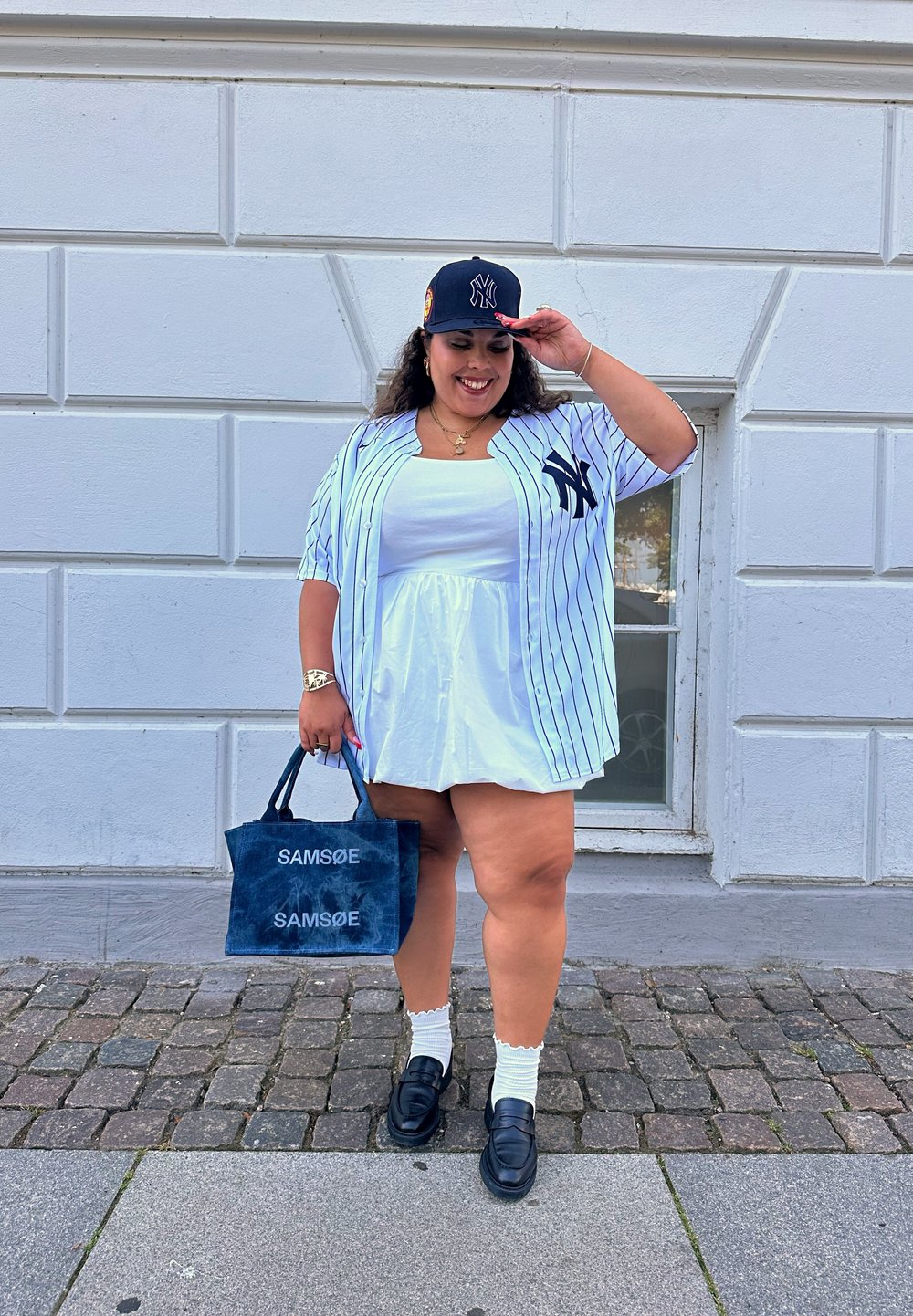 Άσπρο φόρεμα με στενή μέση, μπλε και άσπρο ριγωτό oversized πουκάμισο, μαύρο καπέλο, ναυτικό tote bag με λογότυπα και μαύρες loafers.