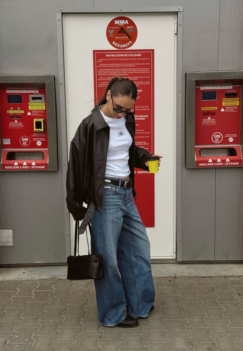 Femme portant des lunettes de soleil, une veste en cuir, une chemise blanche et un jean large tient une tasse jaune et un sac à main noir devant des distributeurs de billets.