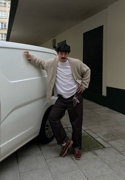 Cárdigan de punto beige, camiseta blanca, pantalones sueltos marrones y zapatos burdeos. Llaves colgando del cinturón. Apoyado contra una furgoneta blanca.