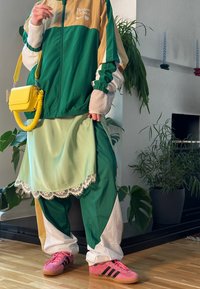 Πράσινη και χρυσή αθλητική ζακέτα με λογότυπο, στρωμένη μίντι φούστα με δαντελένιο τελείωμα, πράσινα και λευκά παντελόνια, ροζ αθλητικά παπούτσια με μαύρες ρίγες, κίτρινη τσάντα.