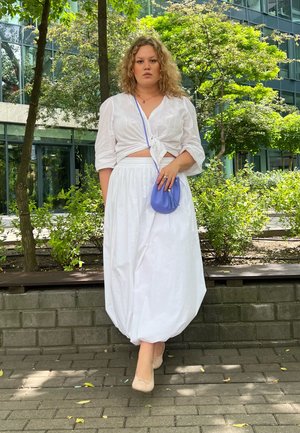 Top corto blanco anudado combinado con una falda larga y fluida. La modelo sostiene un pequeño bolso de mano azul claro. Entorno natural con vegetación.