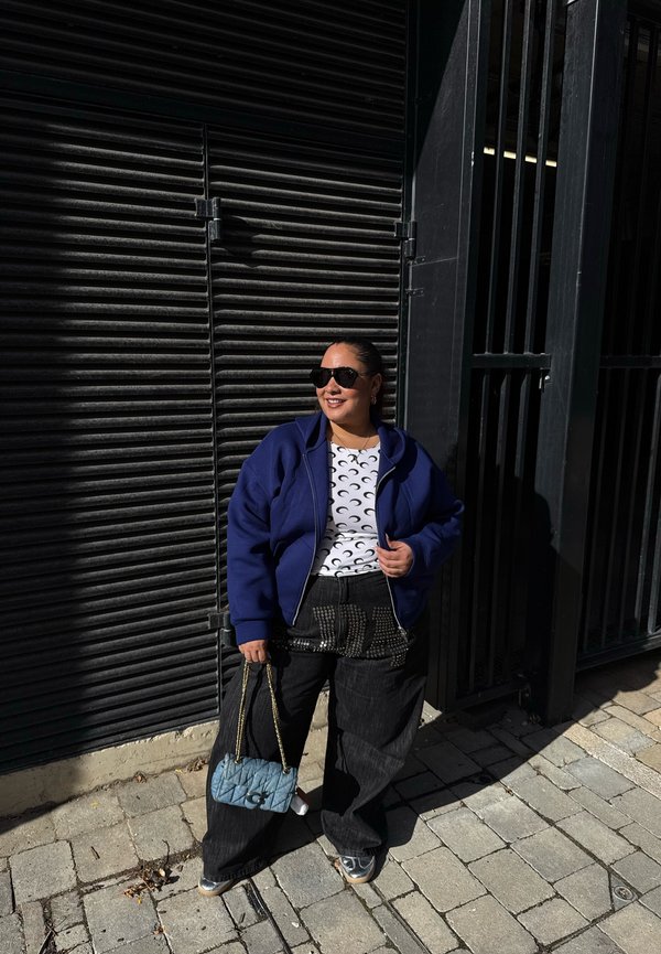 Una mujer con gafas de sol y una chaqueta azul sostiene un bolso azul acolchado junto a una verja de metal negra en una acera soleada.