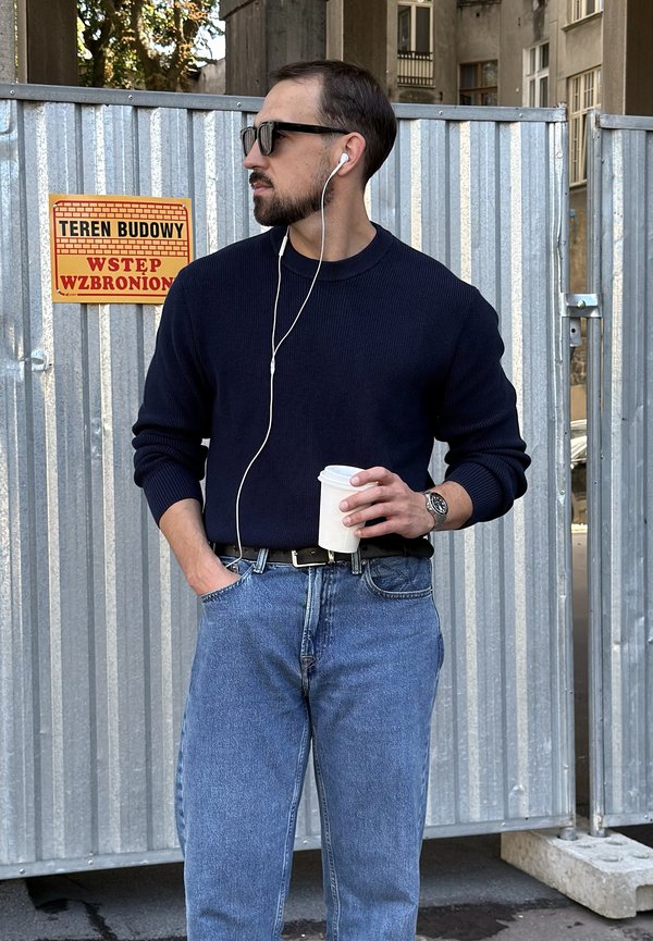 Hombre con gafas de sol y auriculares sostiene una taza de café blanca, de pie frente a una valla metálica con una señal polaca de advertencia de construcción.